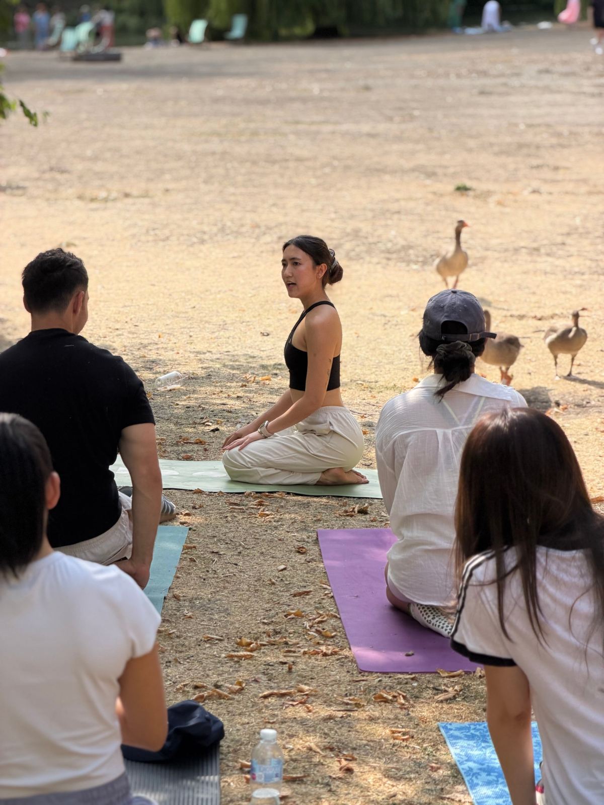 Zu kneeling at the front of an outdoor class, guiding students on their mats