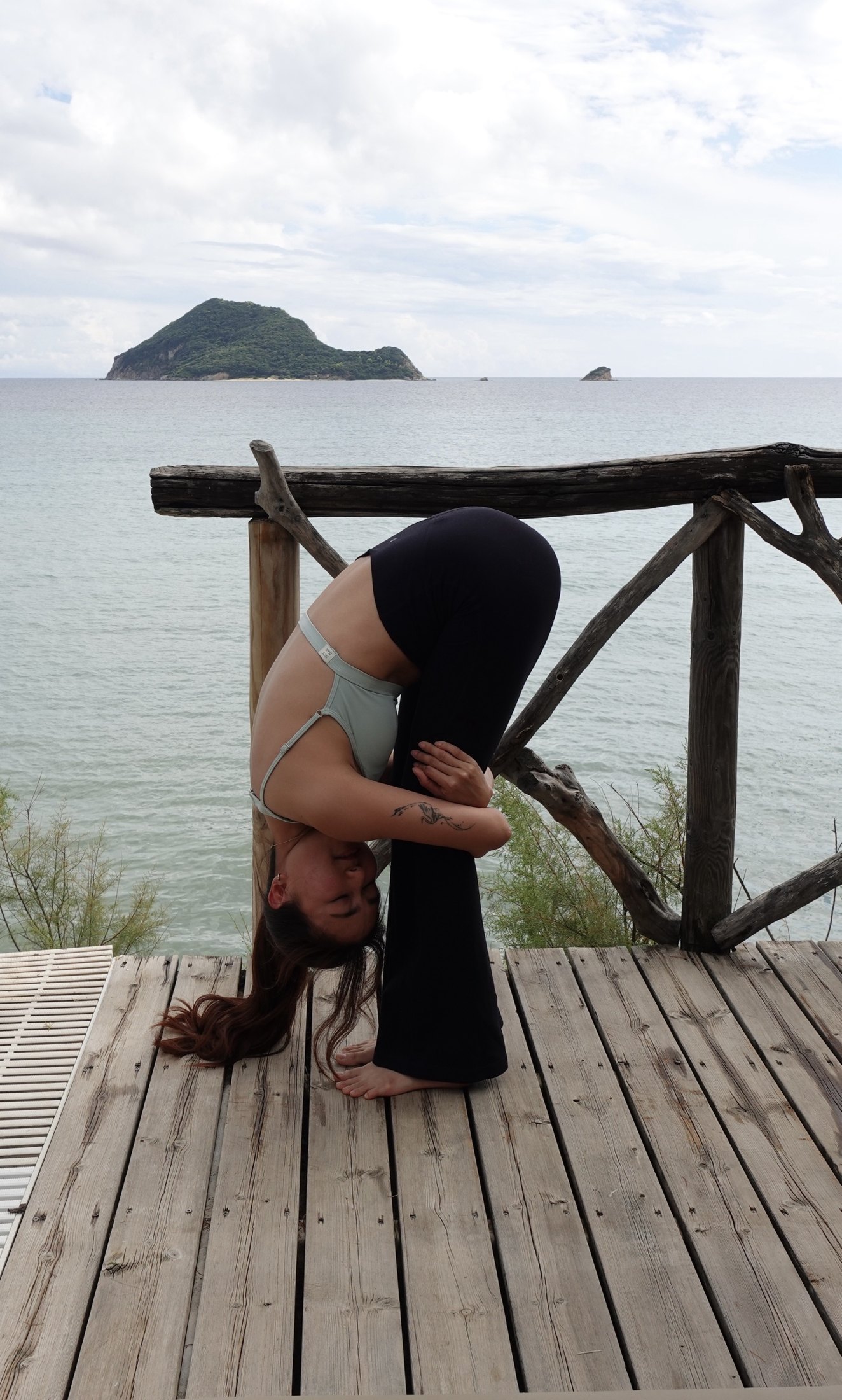 Zu folding forward on a wooden deck above the sea, an island on the horizon behind her